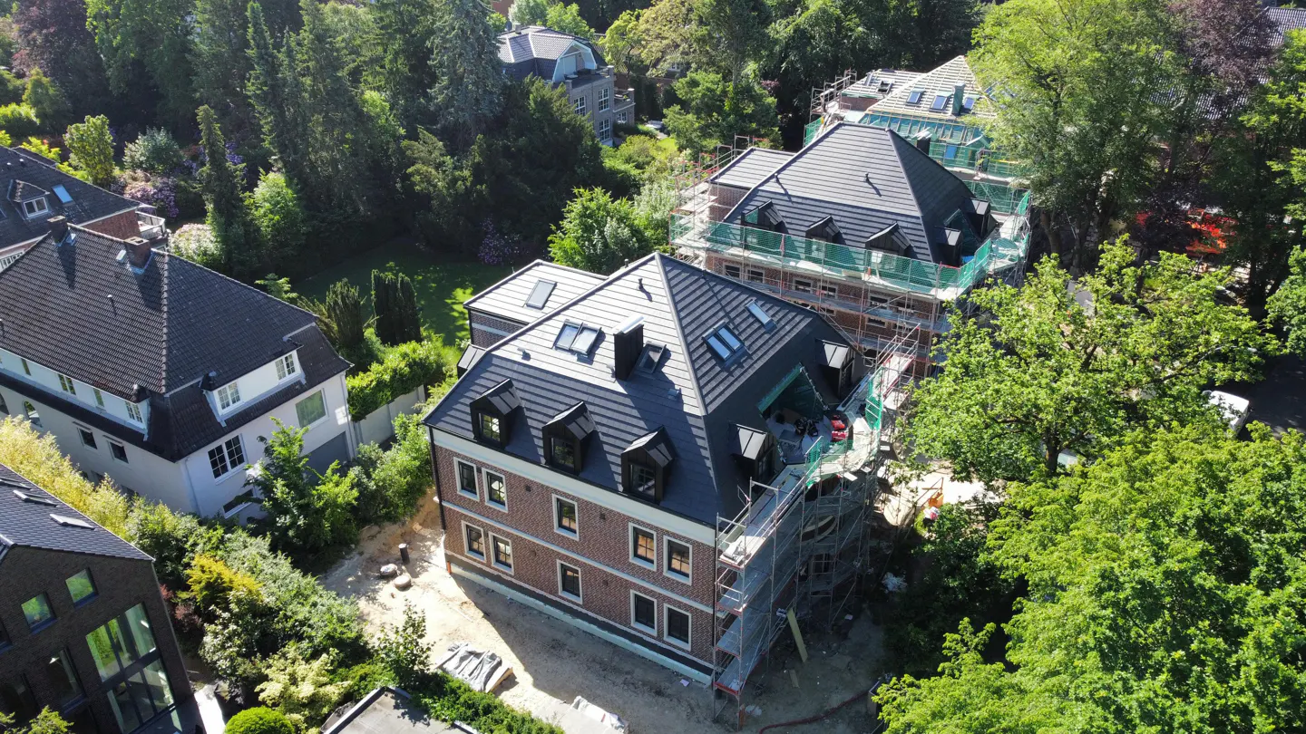 Aerial view of a brick building with a dark roof under construction, surrounded by green trees and other houses.