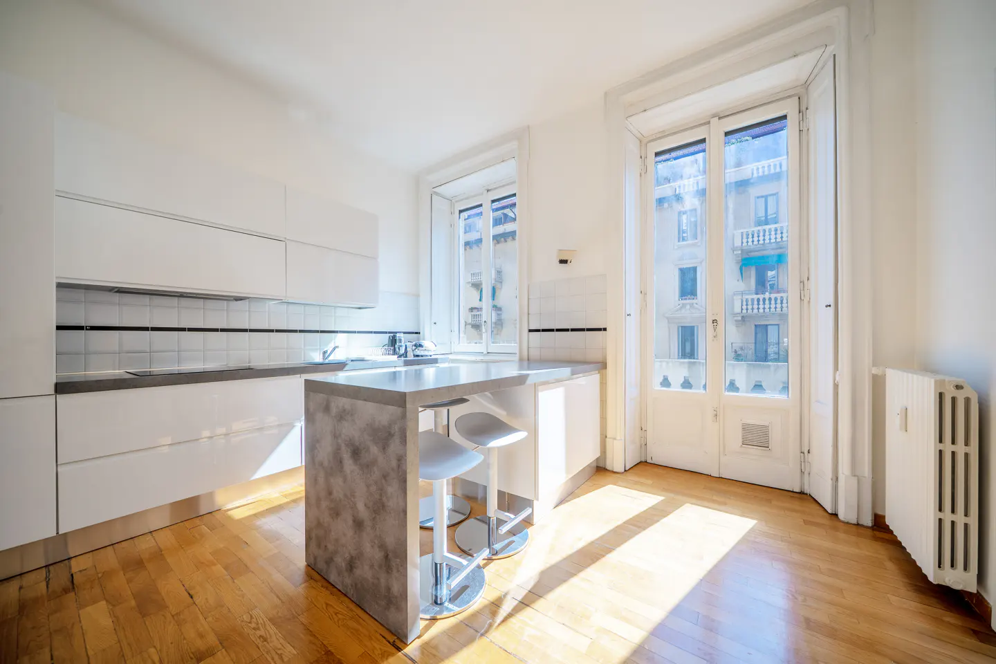 Modern white kitchen with a grey stone island, two bar stools, hardwood floors, and a balcony door overlooking city buildings.