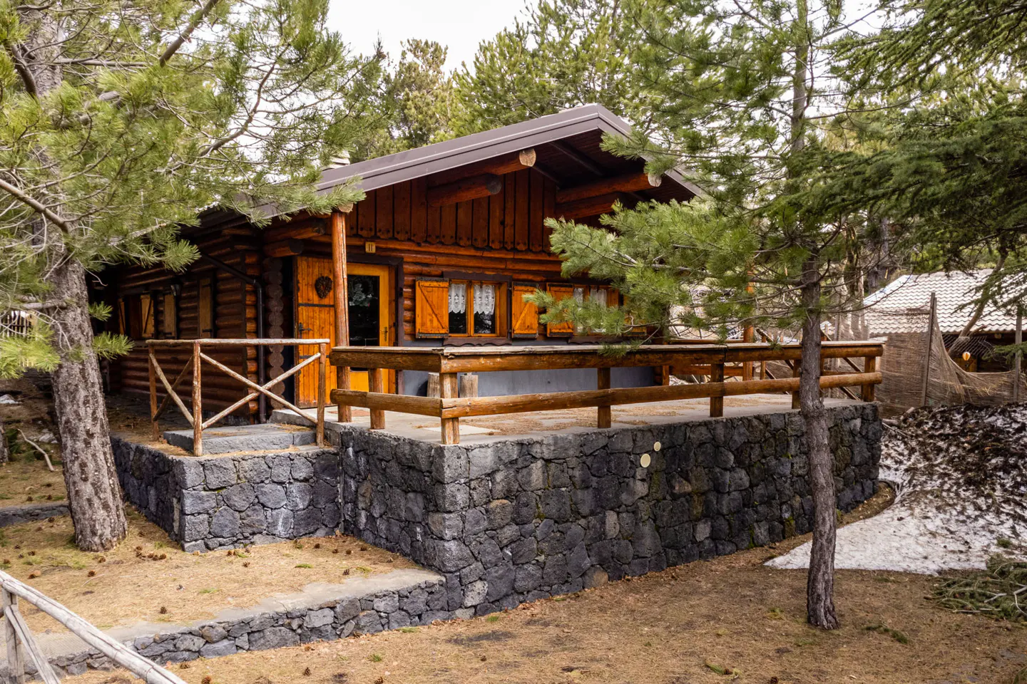 A cozy, two-story log cabin with a stone foundation and wooden railing, surrounded by green pine trees.
