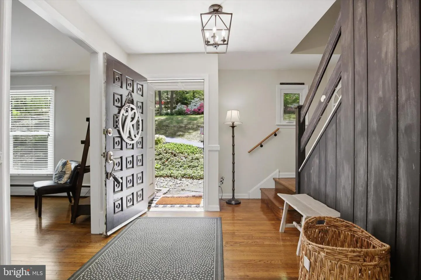 A home's entryway with an open black door, a gray rug, and a wooden staircase. A lantern hangs from the ceiling.