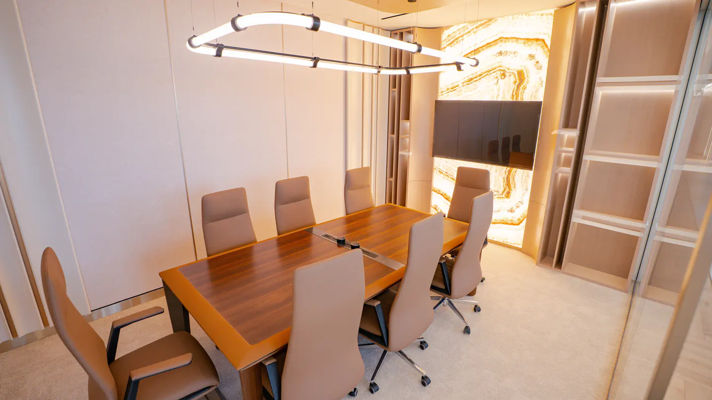 Conference room with a wood table, tan chairs, and a modern light fixture. A TV is mounted on a marble wall.