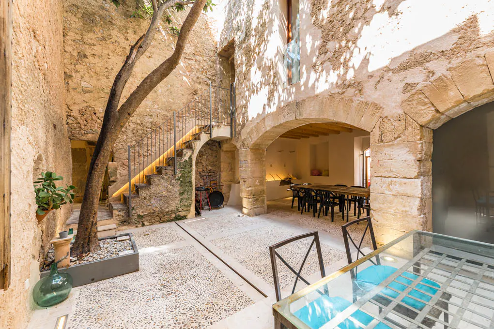 Stone courtyard with stairs, tree, and dining area. Arched doorways lead to interior spaces. Table with blue cushions in foreground.
