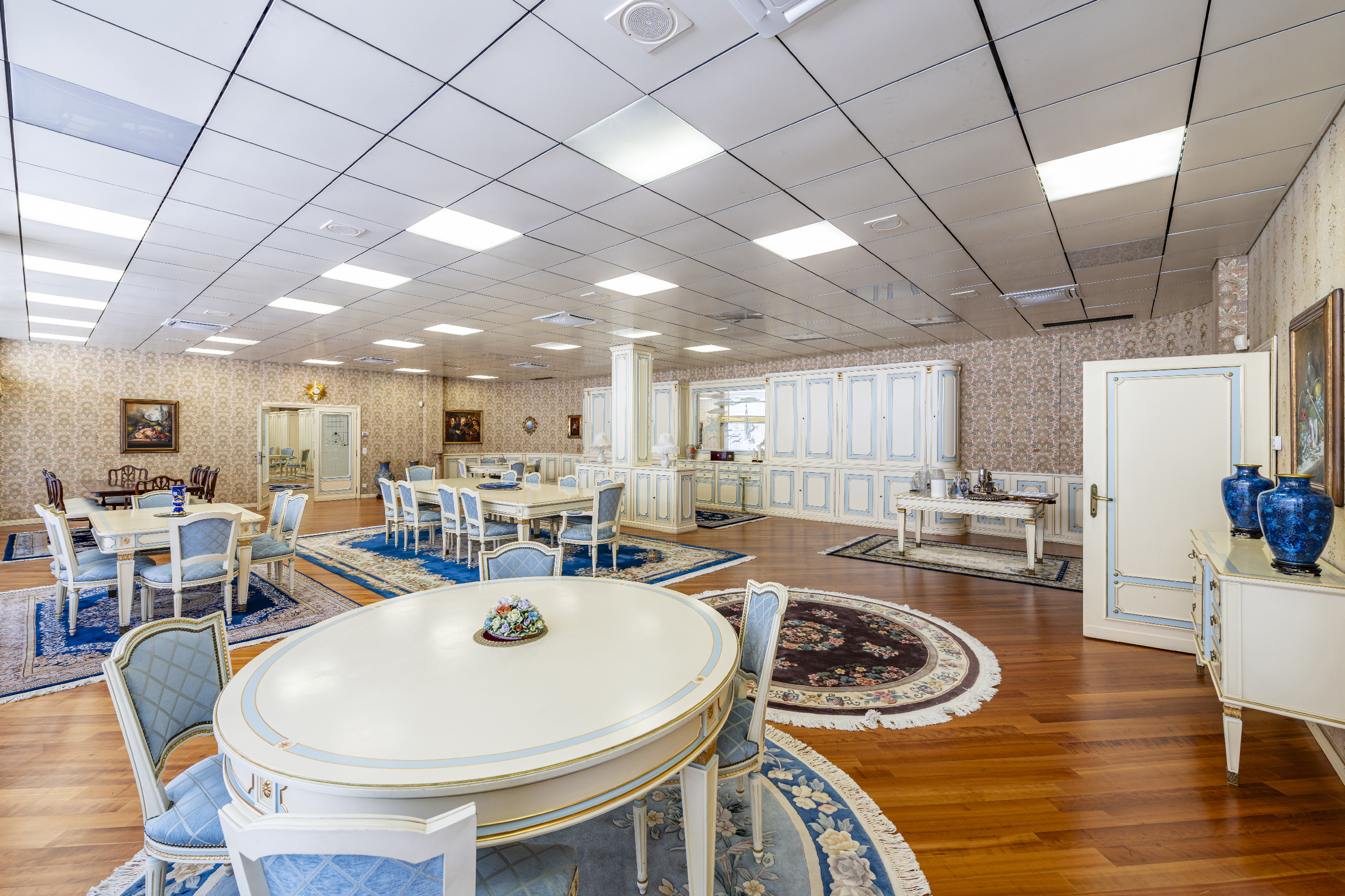 Spacious dining room with multiple white tables, blue upholstered chairs, patterned rugs, and wood floors under a grid ceiling. [AI-generated text]