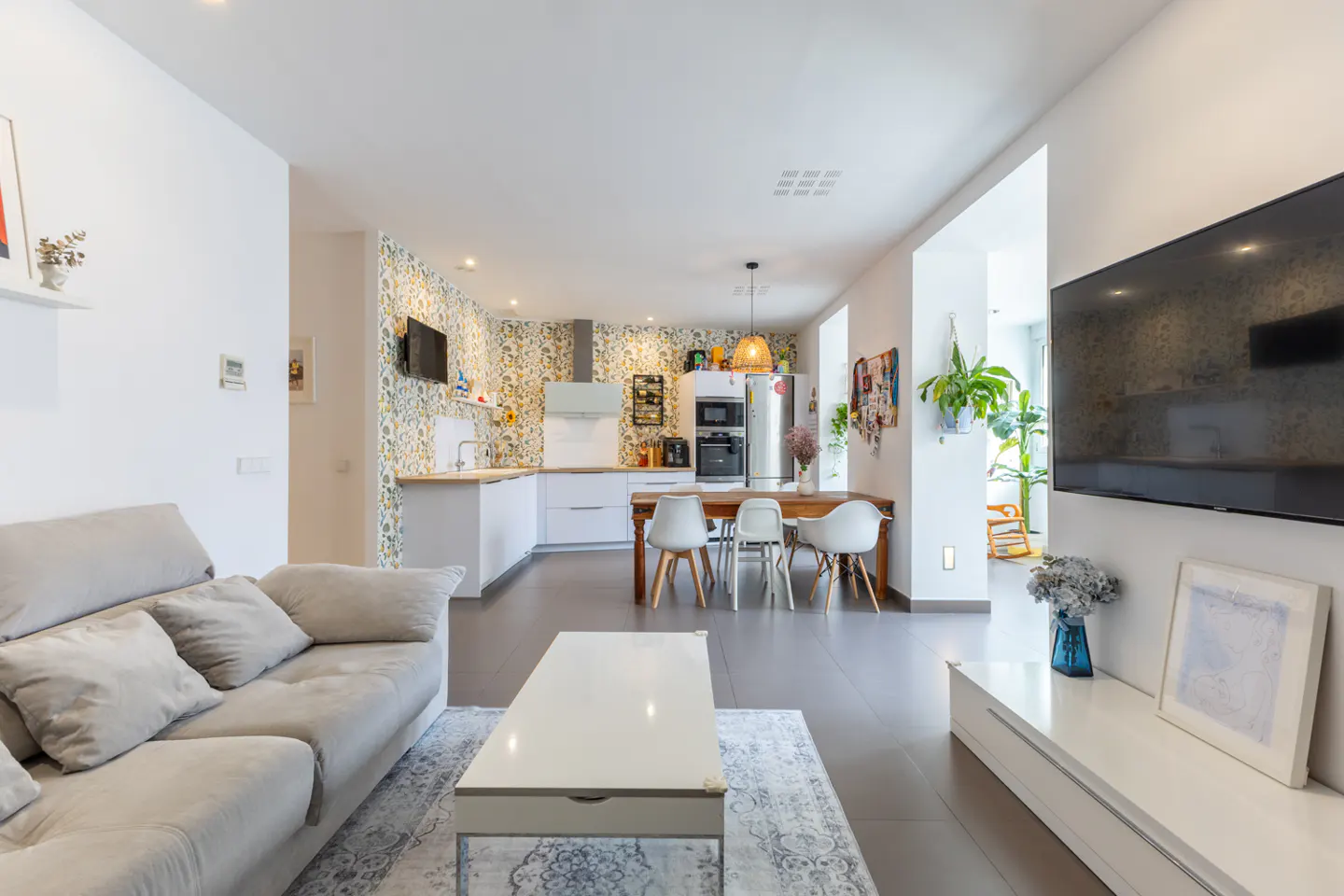 Open-concept living space with a gray sofa, white coffee table, and a kitchen with floral wallpaper in the background.