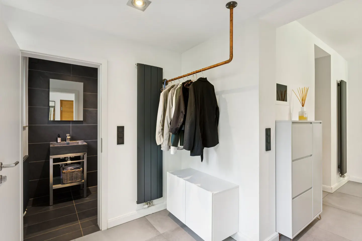 Hallway with a copper pipe clothing rack holding jackets, a white cabinet, and a view into a black-tiled bathroom.