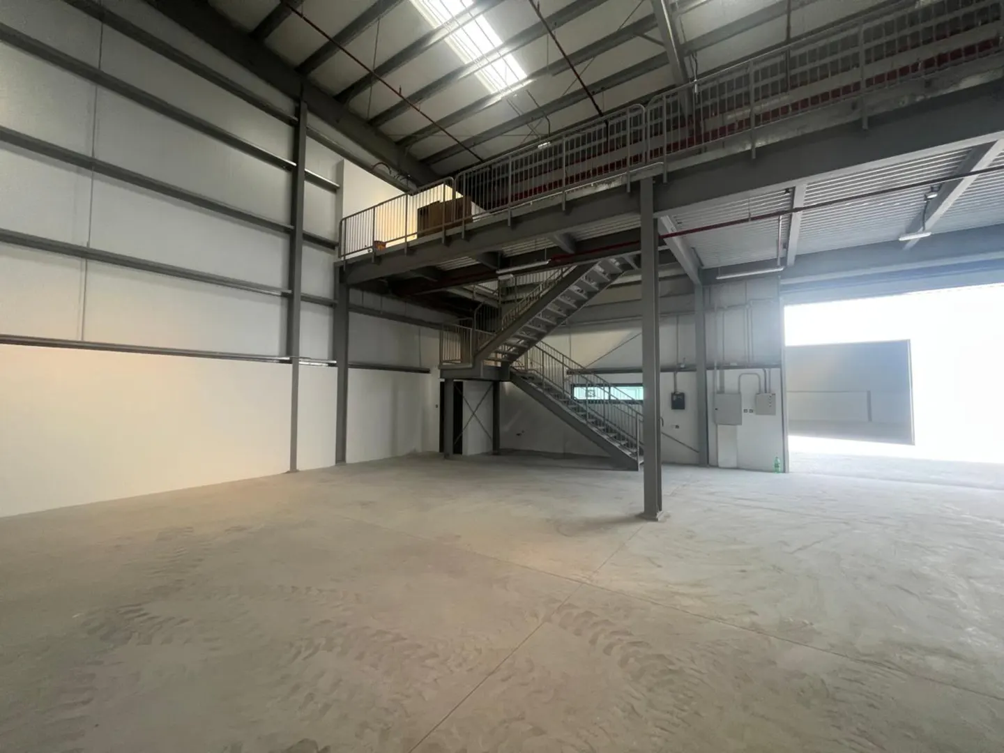An empty warehouse interior with a metal mezzanine and stairs. The walls are white, and the floor is concrete. A large door is open, letting in light.