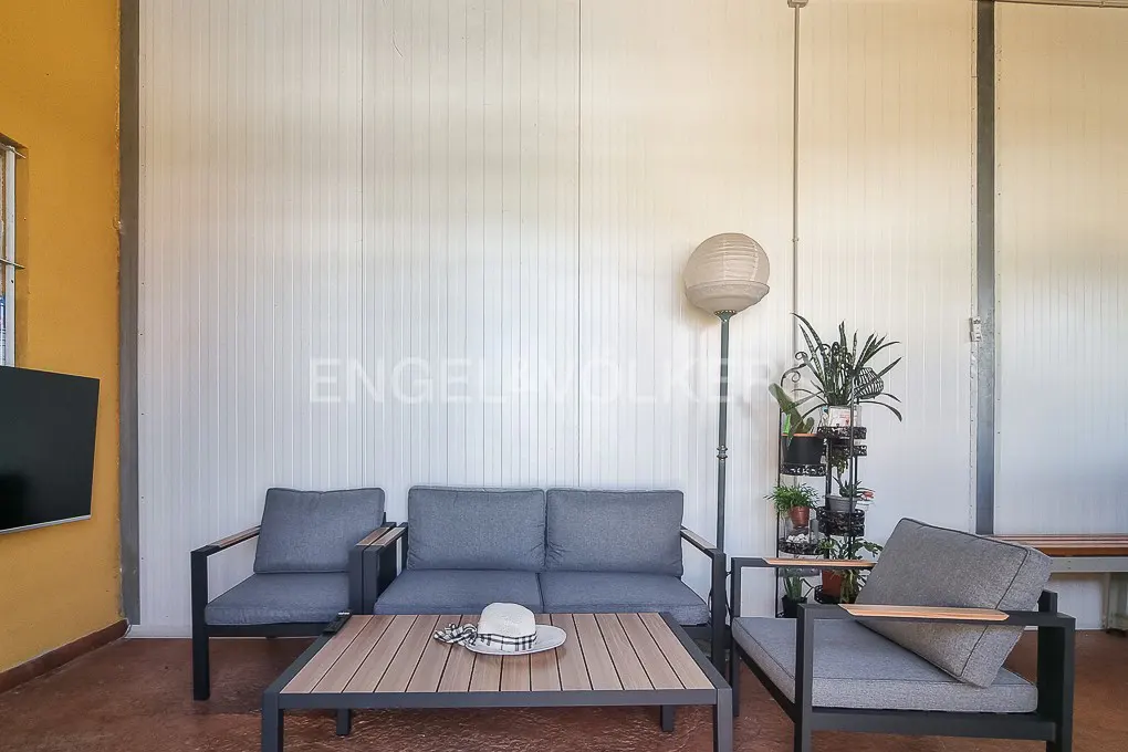 Outdoor living space with gray cushioned seating, a wood table with a hat, and potted plants against a white paneled wall.