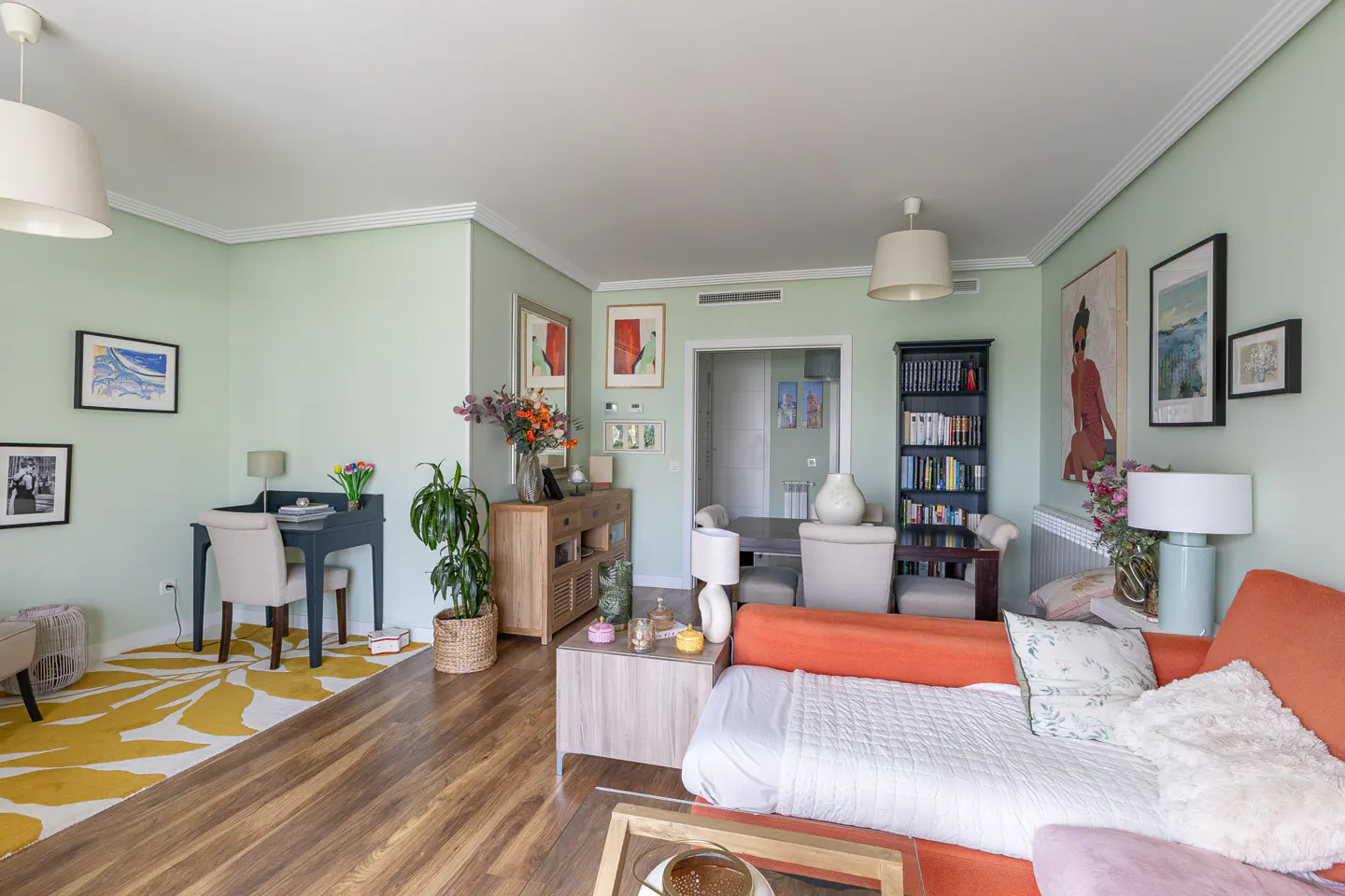 Bright living room with sage green walls, orange sofa, yellow patterned rug, wooden furniture, and framed wall art.
