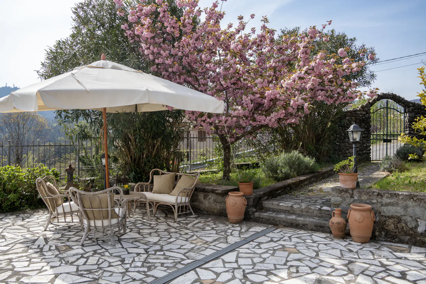 Outdoor patio with stone tiles, wicker furniture under a white umbrella, and a blooming pink tree. Stone archway in the background.