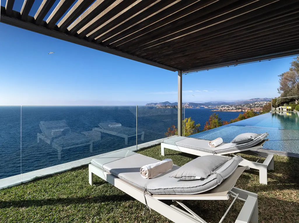 Ocean view patio with two lounge chairs on green grass, under a pergola. An infinity pool is to the right, with a view of the ocean and distant land.