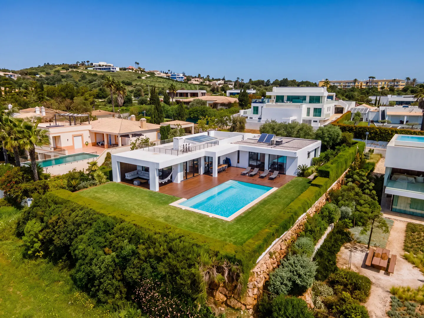 Aerial view of a modern white house with a pool, surrounded by green lawns and trees under a clear blue sky.