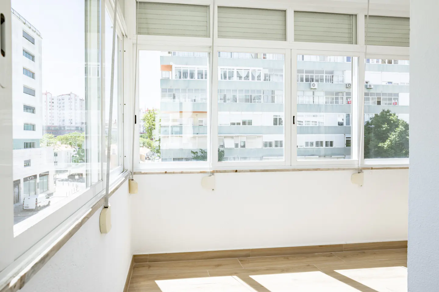 Bright, empty sunroom with white walls, wood-look tile floors, and large windows overlooking a city street and buildings.