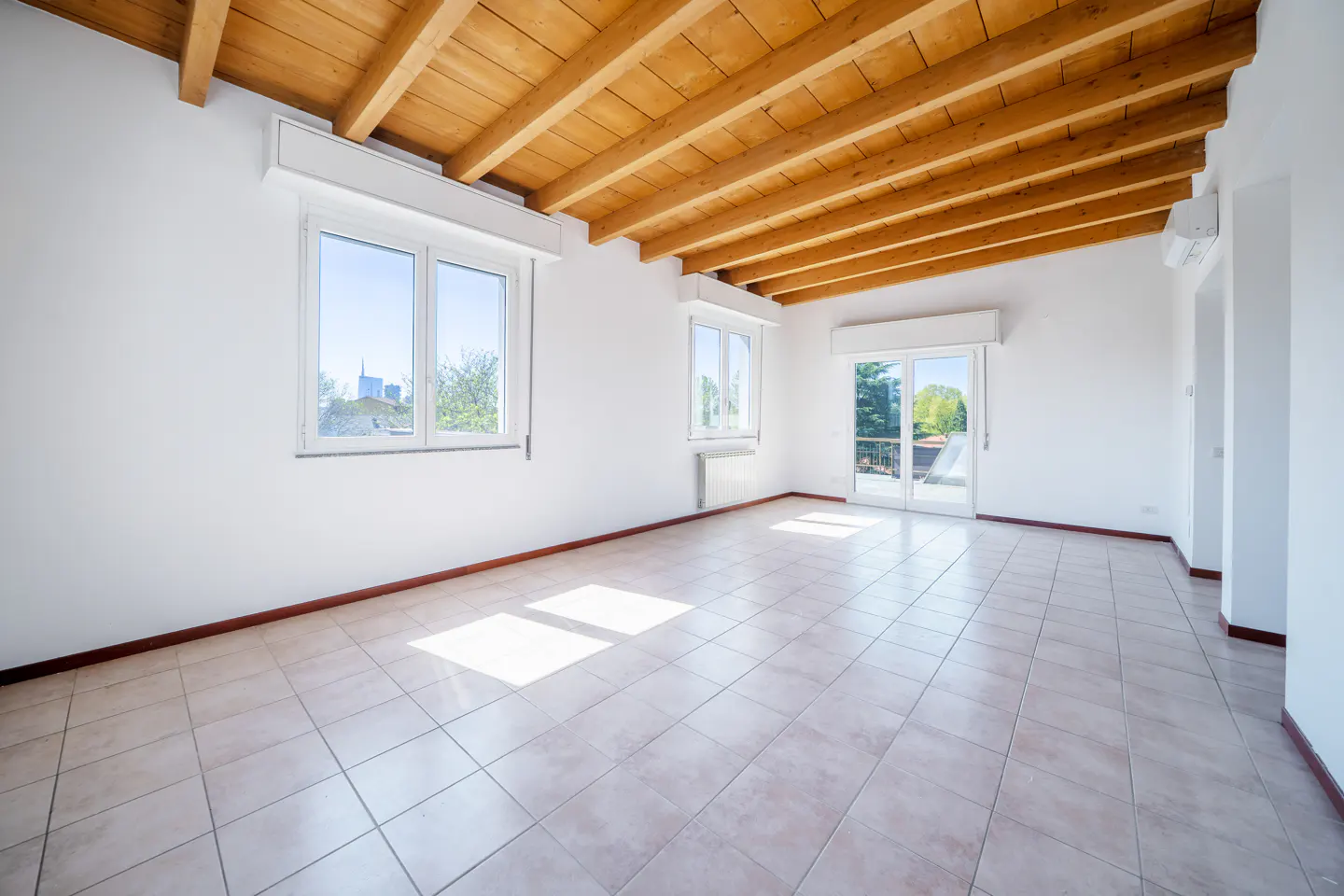 Bright, empty room with white walls, tile floor, and exposed wood beam ceiling. Windows offer a city view.