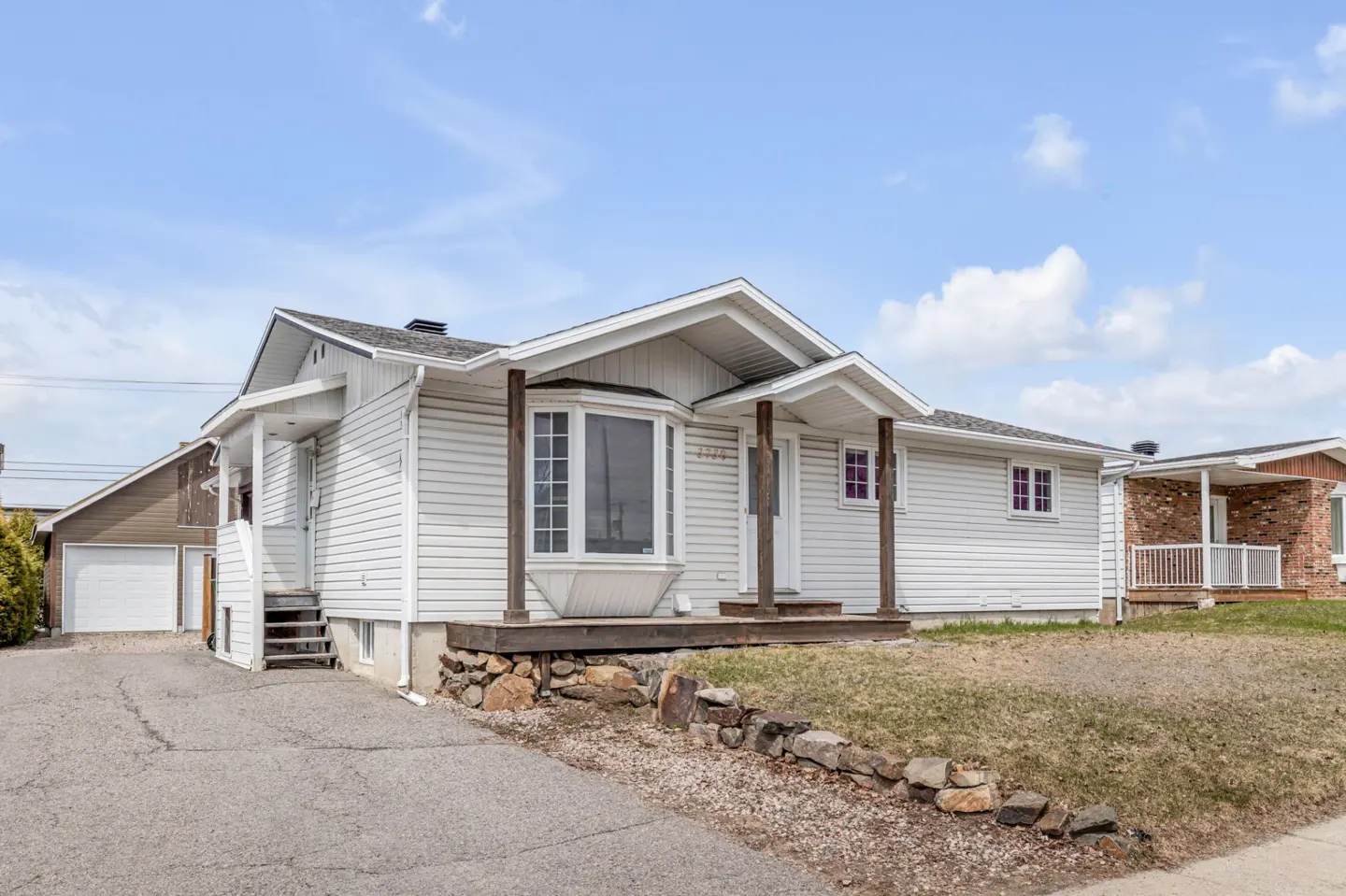 Exterior view of a one-story white house with a bay window and a small porch with wooden pillars. A detached garage is visible on the left.