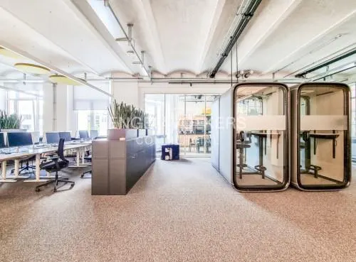 Bright office space with desks, chairs, plants, and two phone booths. The walls are white and the floor is beige.