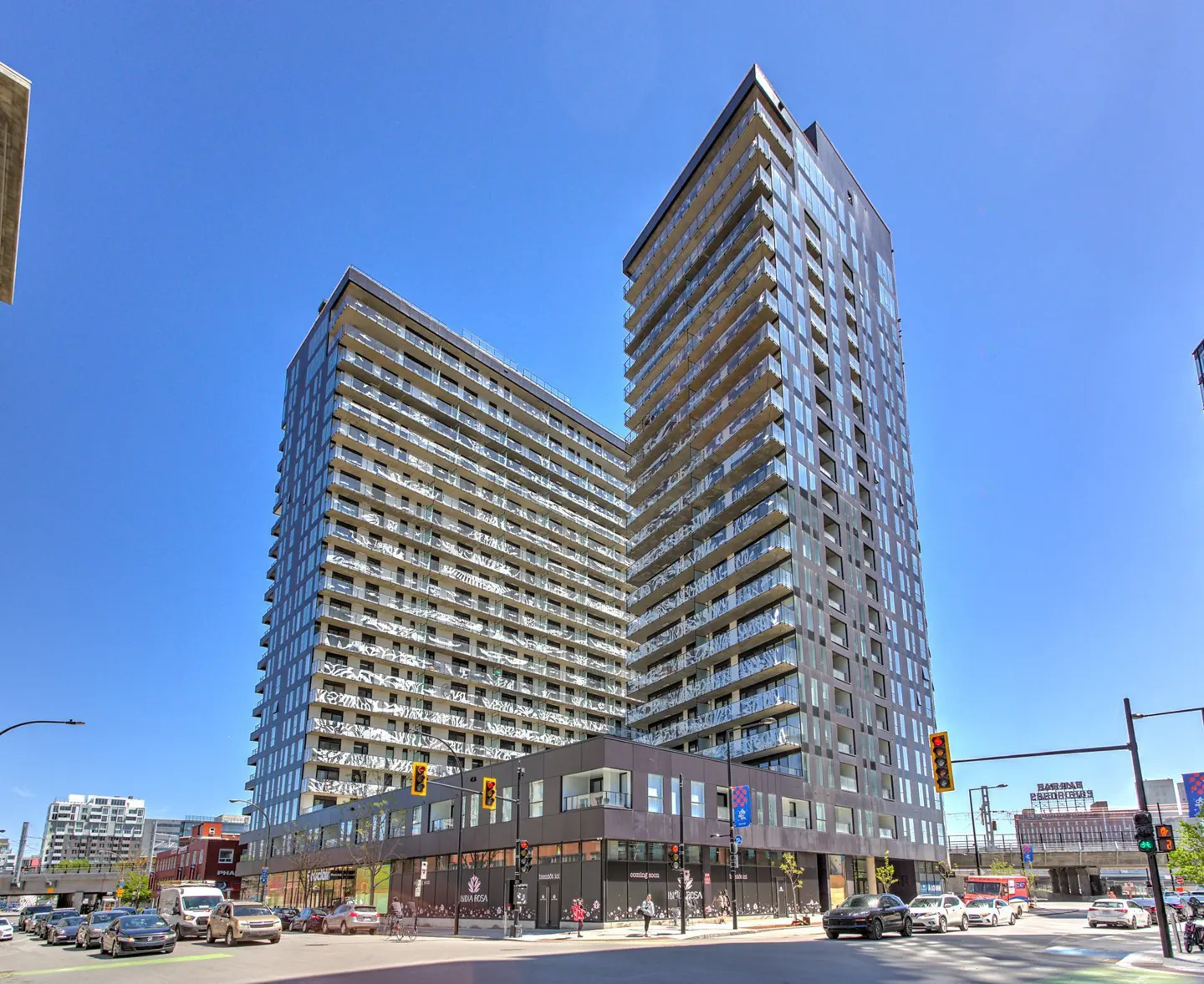 Two modern high-rise apartment buildings with glass balconies under a clear blue sky. Street-level retail and traffic are visible.