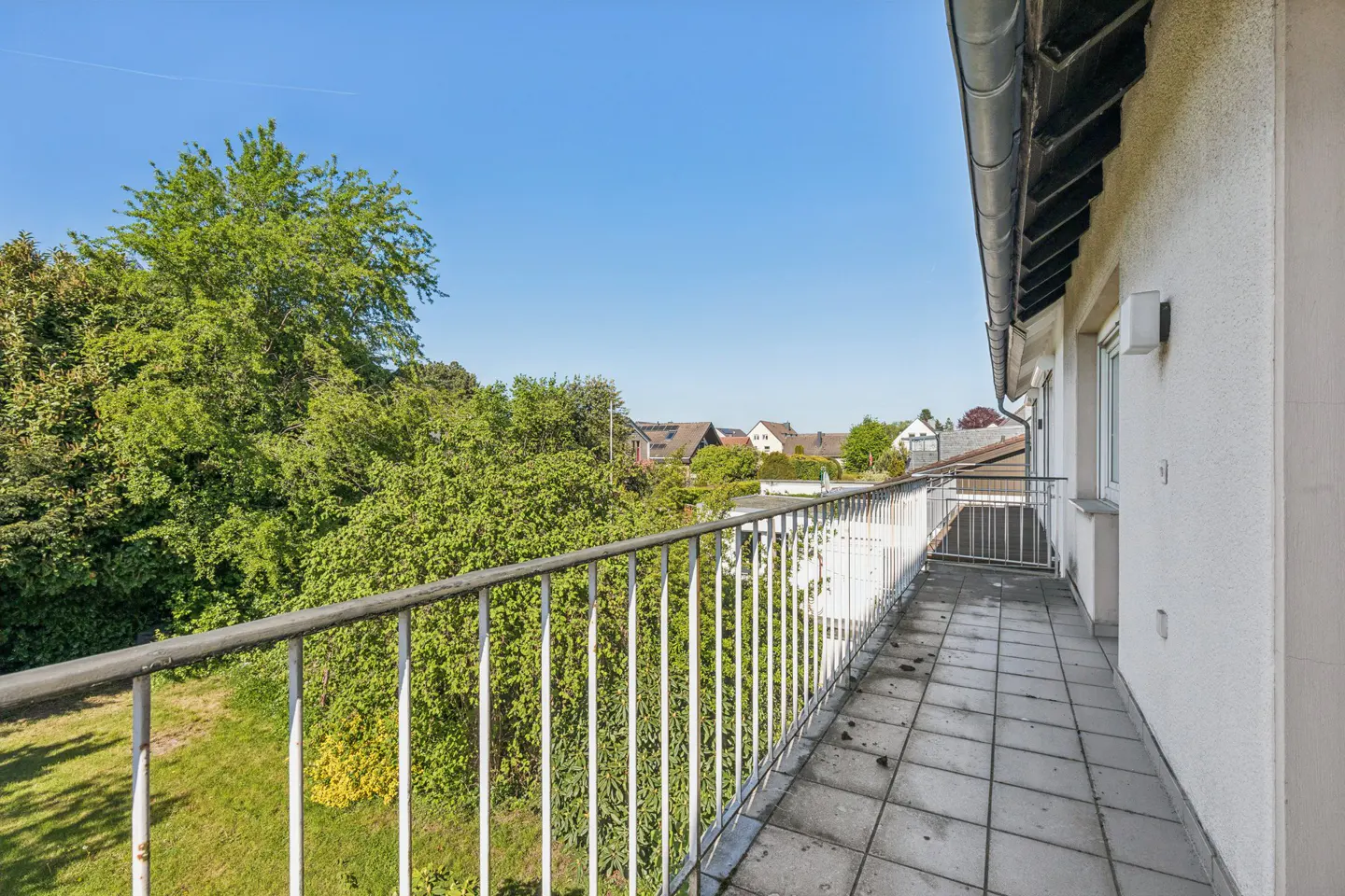 Balcony with white railings and gray tiled floor overlooks green trees and houses under a blue sky.
