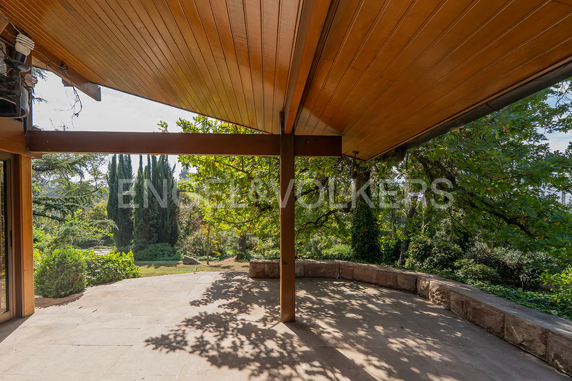 Covered patio with a wood ceiling and stone floor, looking out onto a lush green garden with trees and shrubs.