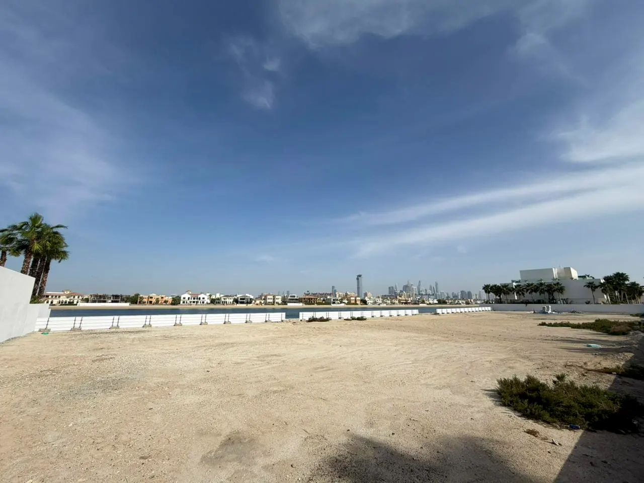 Vacant sandy lot with water view. Palm trees, houses, and city skyline in the background under a blue sky.