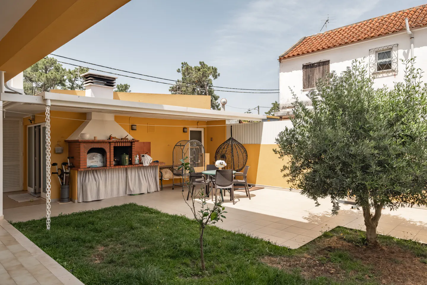 Outdoor patio with a brick grill, dining table, and hanging chair. A green lawn and olive tree add to the ambiance.
