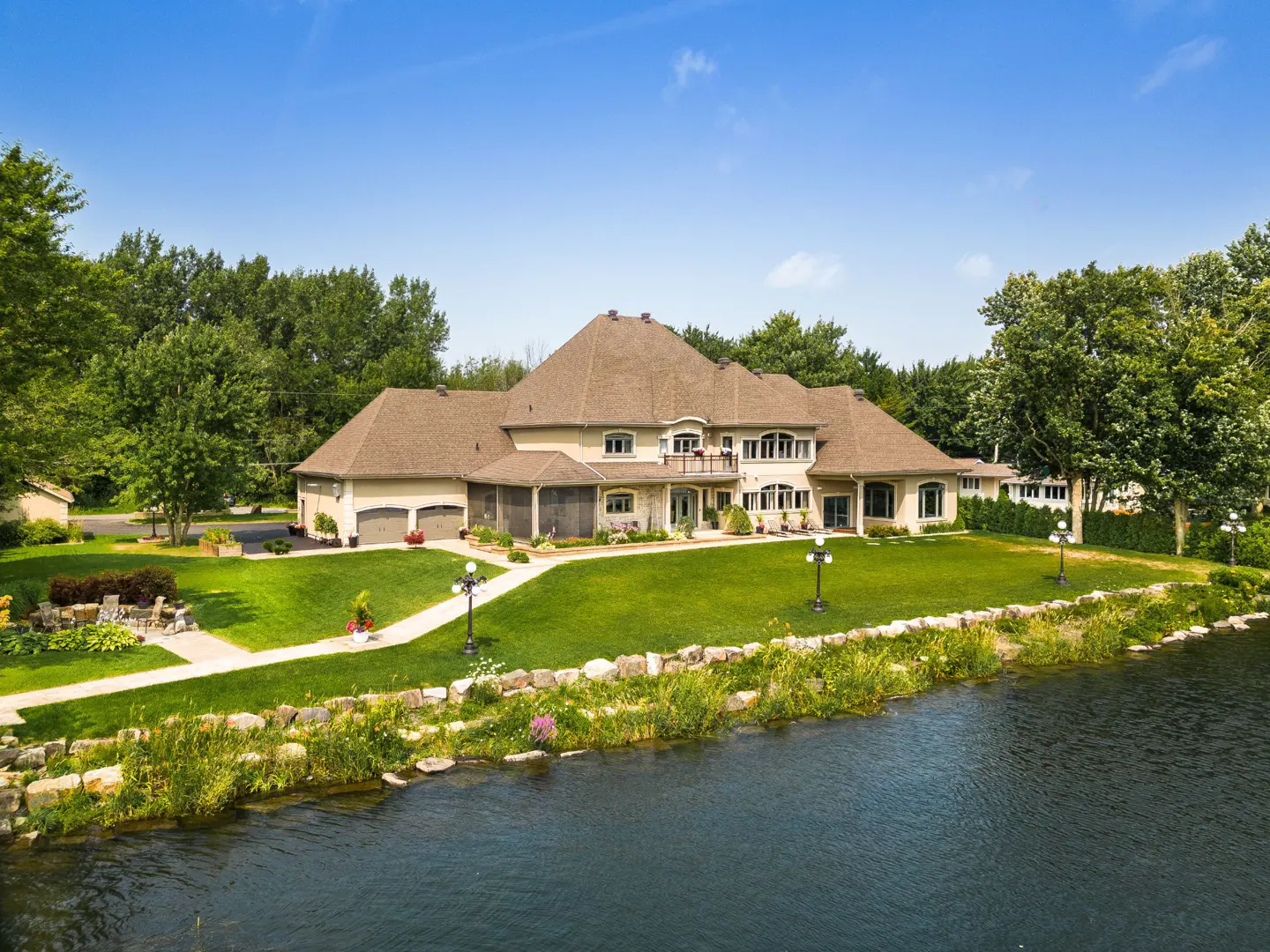 Aerial view of a large beige house with a brown roof, green lawn, and a pond in front. Blue sky above.