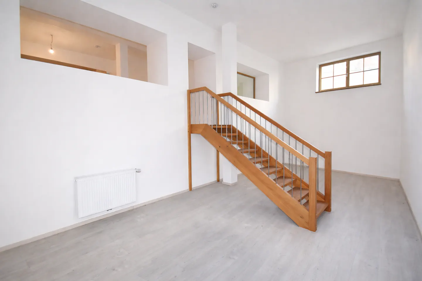 Bright, empty room with a wooden staircase and gray floors. White walls feature a window and cut-out spaces. A radiator is on the left wall.