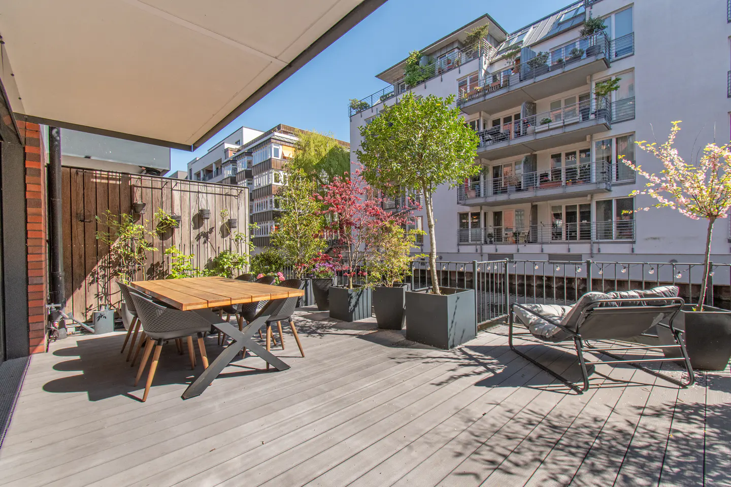A sunny patio with a wooden table, chairs, and a lounge chair. Potted trees line the railing, with buildings in the background.