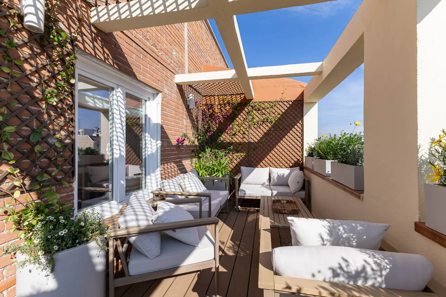 A sunlit rooftop patio with wooden furniture, white cushions, and potted plants. A brick wall and trellis provide privacy.