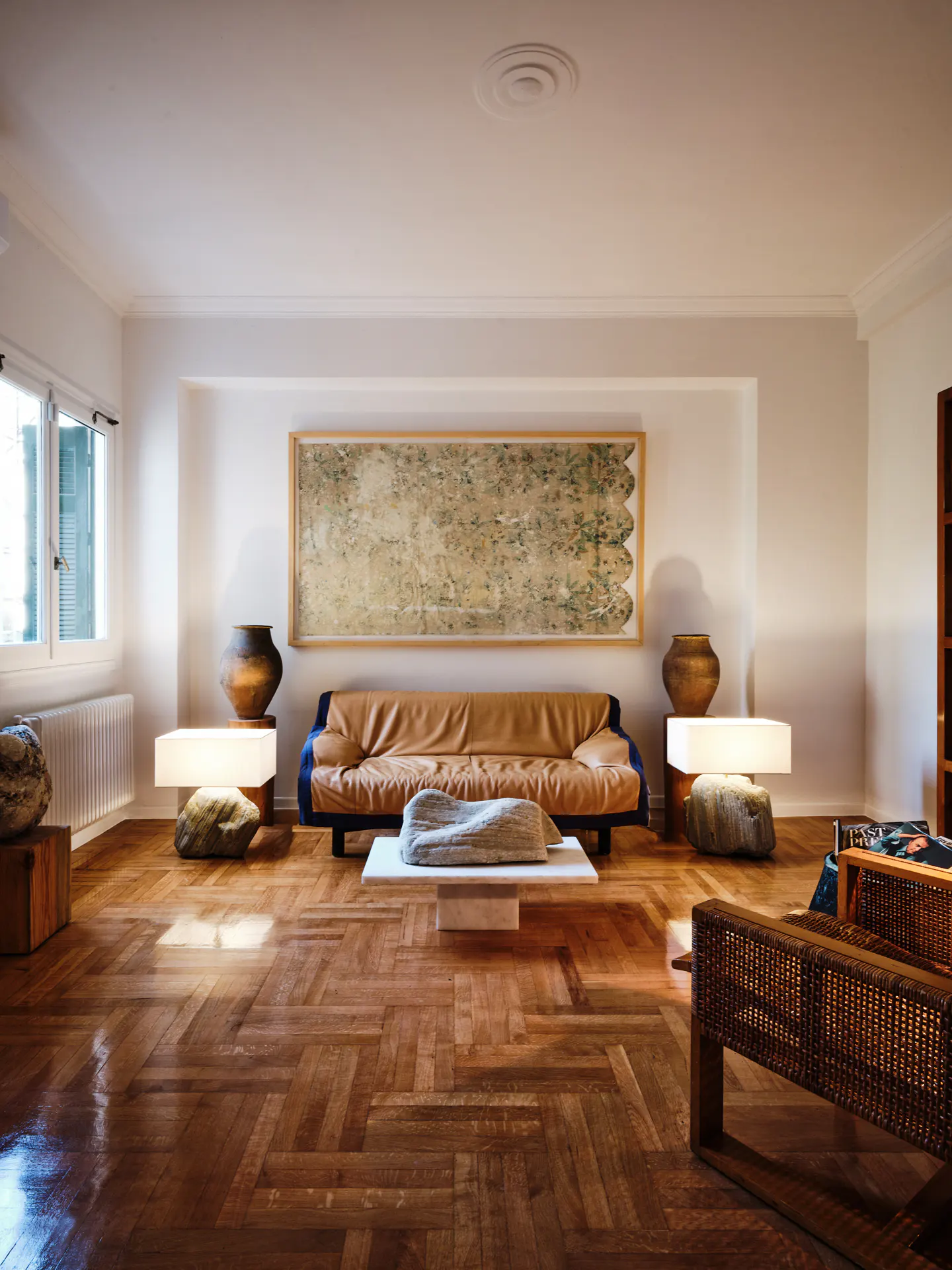 Bright living room with herringbone wood floors, tan leather couch, and a large framed painting above. Lamps sit on rock bases.