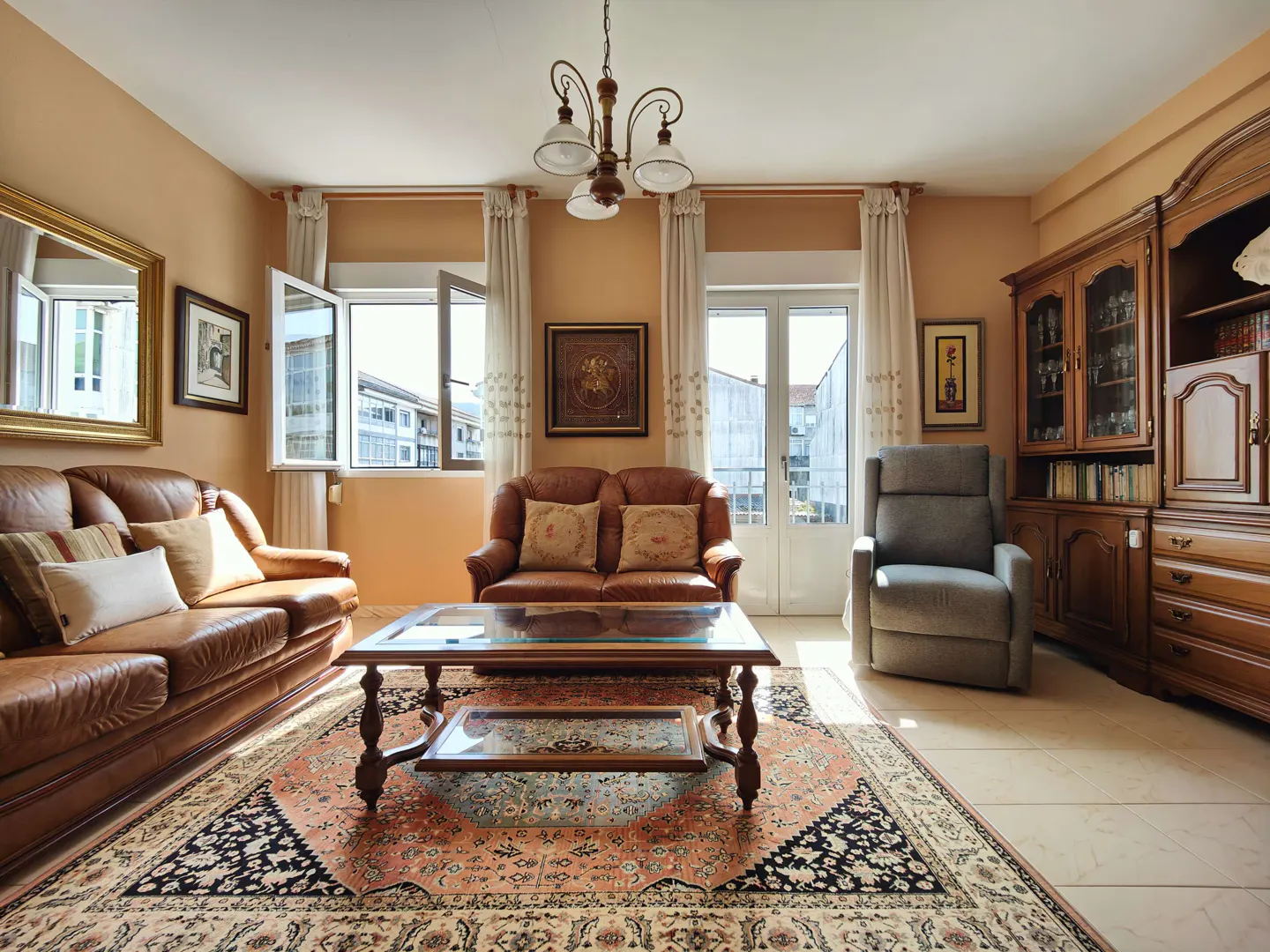 Living room with brown leather sofas, a glass coffee table on an ornate rug, and a wooden cabinet. Natural light streams through open windows.