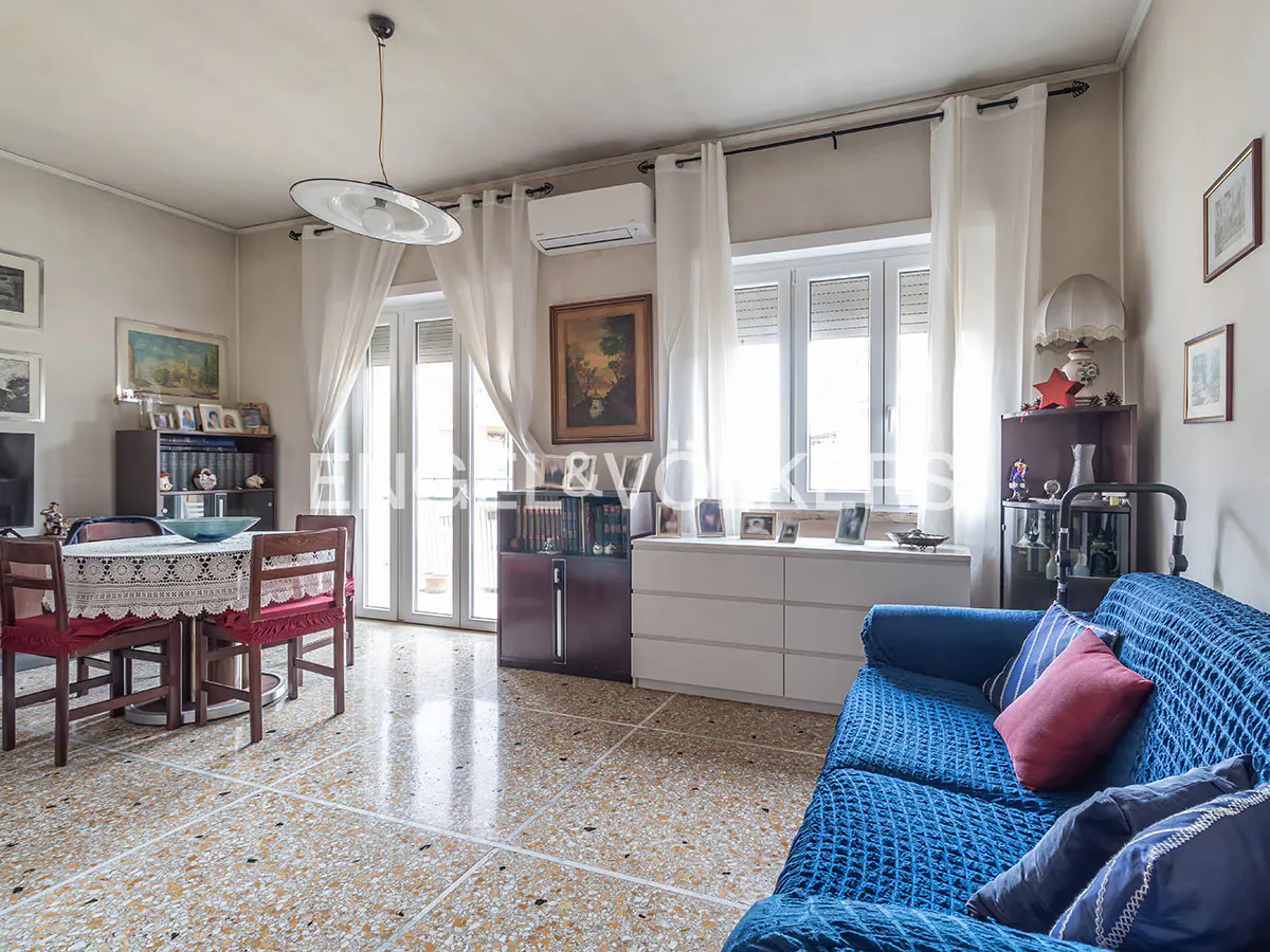 A bright living room with a blue sofa, dining table, and white curtains. The floor is tiled with a terrazzo pattern.
