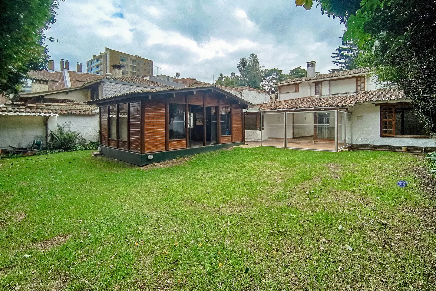 Backyard view of a white house with a brown-tiled roof, a wooden cabin, and a green lawn on a cloudy day.