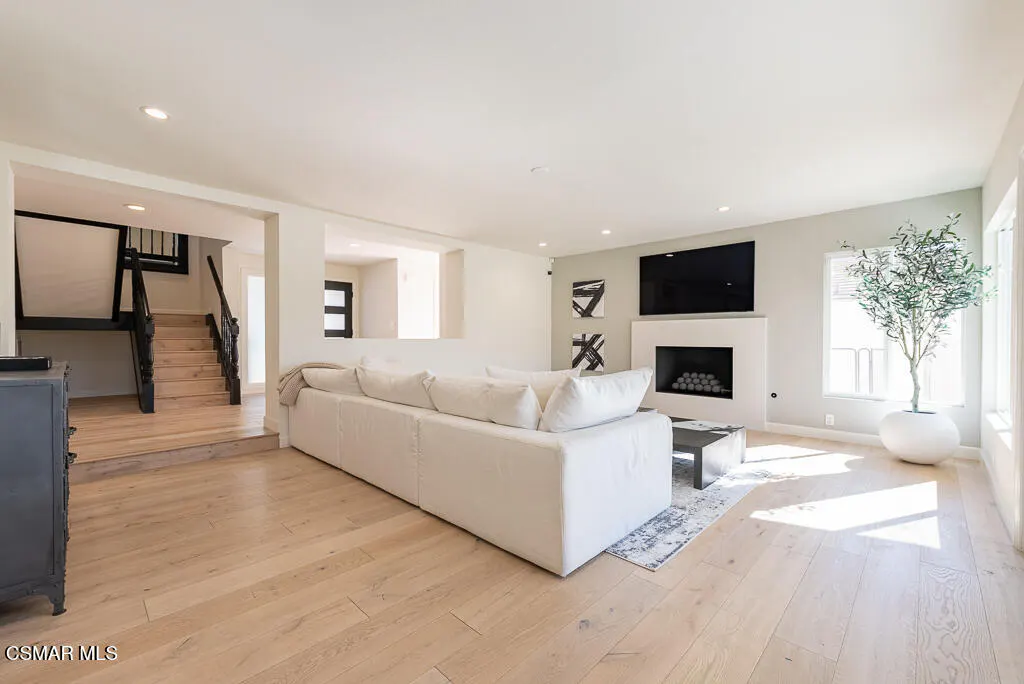 Modern living room with light wood floors, white sectional sofa, fireplace with mounted TV, and a potted olive tree.