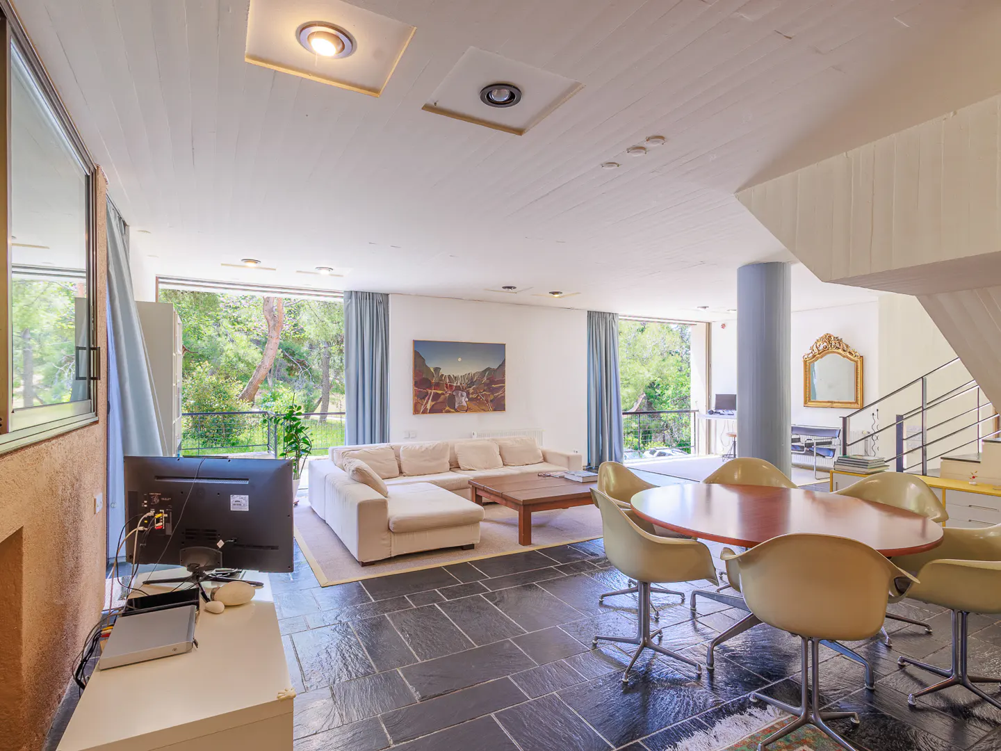 Open-concept living room with slate floors, white walls, and large windows overlooking greenery. A white sofa, wood table, and dining set furnish the space.