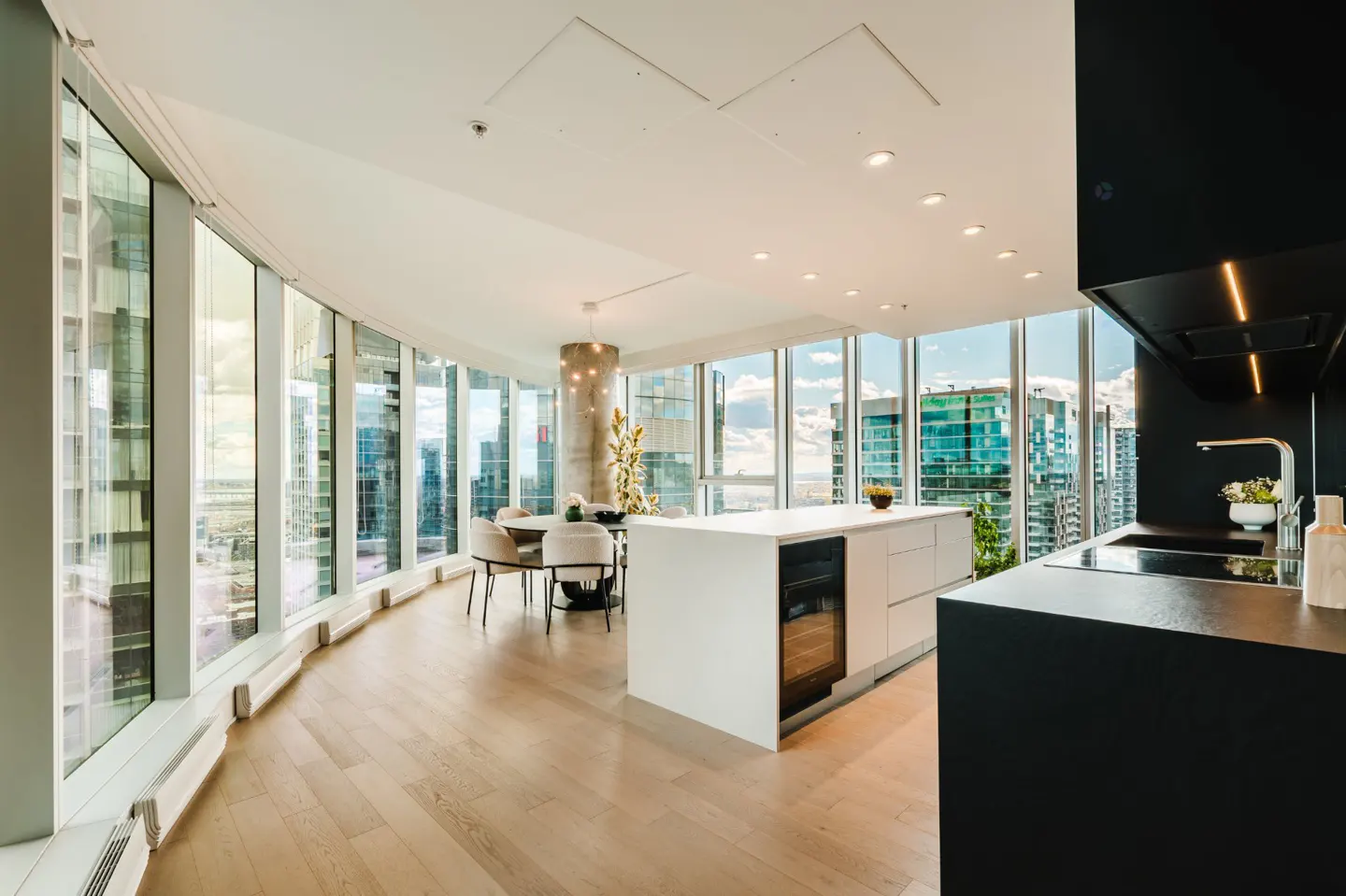 Bright, modern condo interior with floor-to-ceiling windows offering a city view. Features a white kitchen island, black countertops, and a dining area.