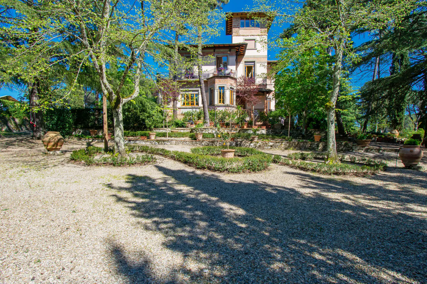 Exterior view of a multi-story beige house with a tower, surrounded by trees and a gravel driveway.