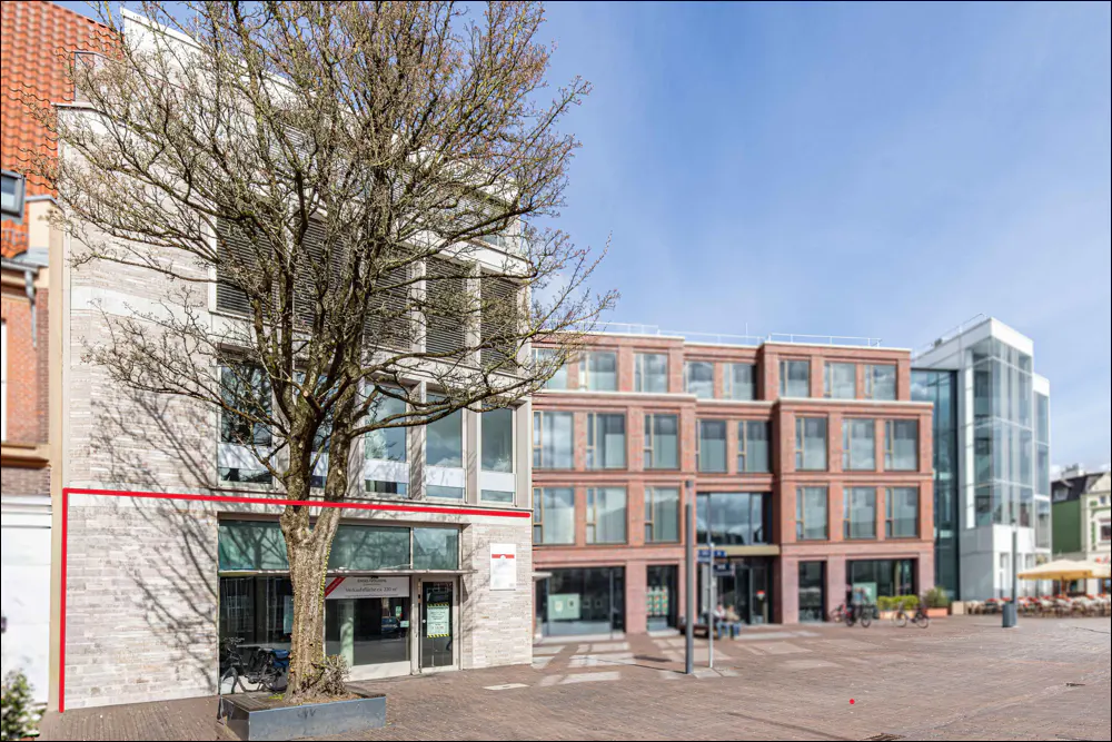 Exterior view of a modern, multi-story building with a light gray brick facade and large windows, framed by a bare tree in the foreground.