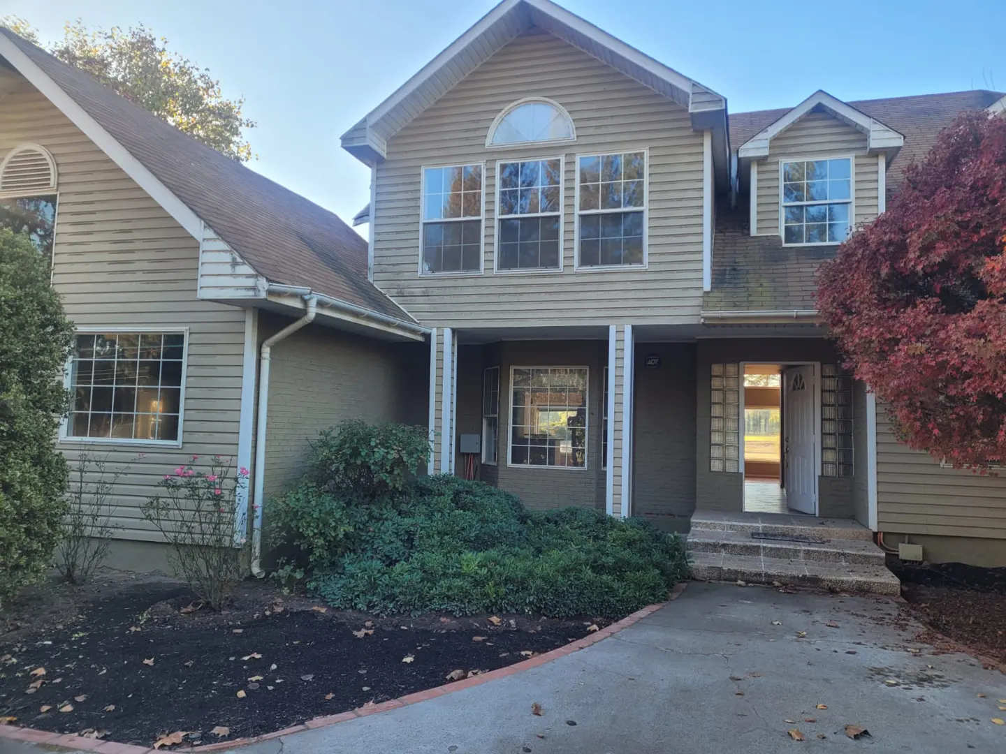 Two-story beige house with a brown roof, green bushes, and a red tree on a sunny day. The front door is open.