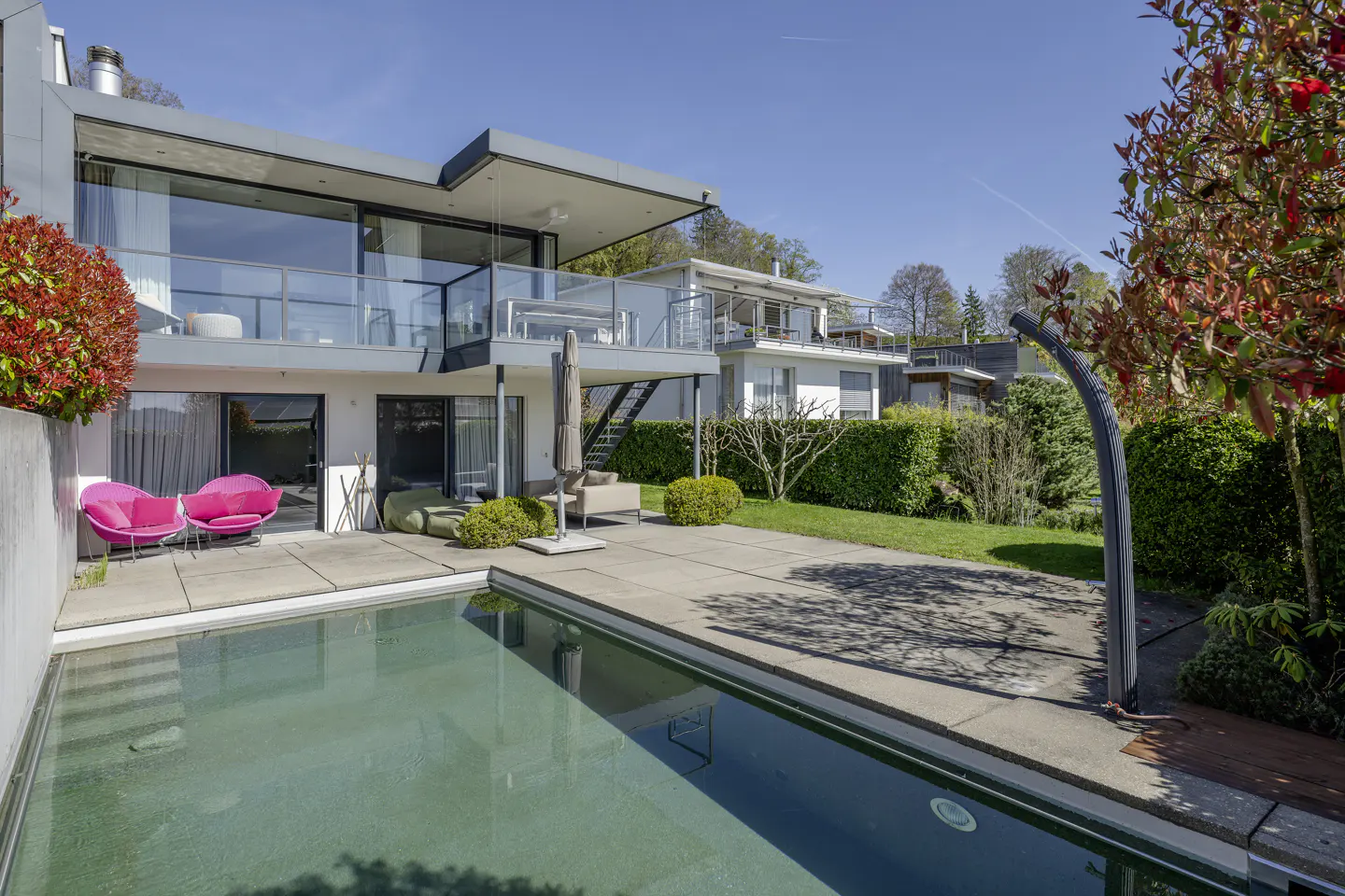 Modern two-story house with a pool. Pink chairs sit on the patio. Green lawn and trees surround the property. Blue sky.