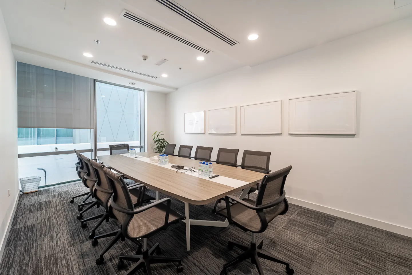 Bright, modern conference room with a long wooden table, black chairs, and white walls. Large window with blinds and whiteboards on the wall.