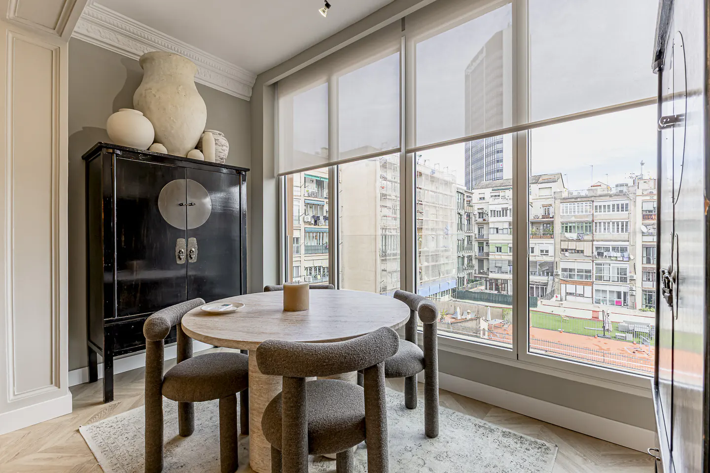 A dining area with a round table, four gray chairs, and a black cabinet with vases. Large windows overlook a city.