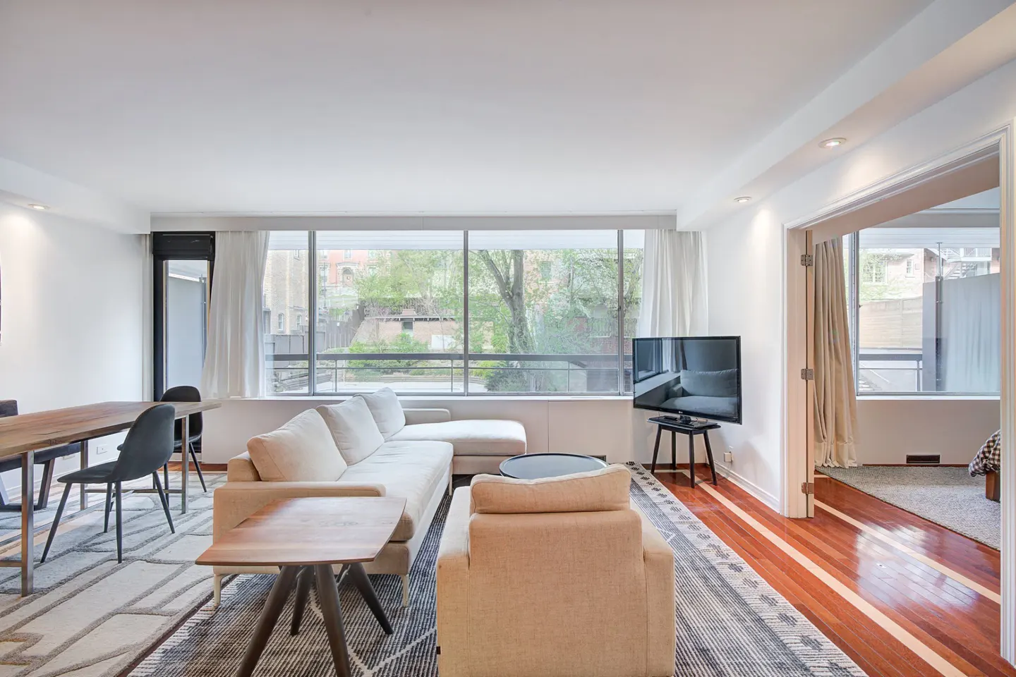 Bright living room with a beige sectional sofa, wood dining table, and large windows overlooking greenery. Hardwood floors and a patterned rug add warmth.