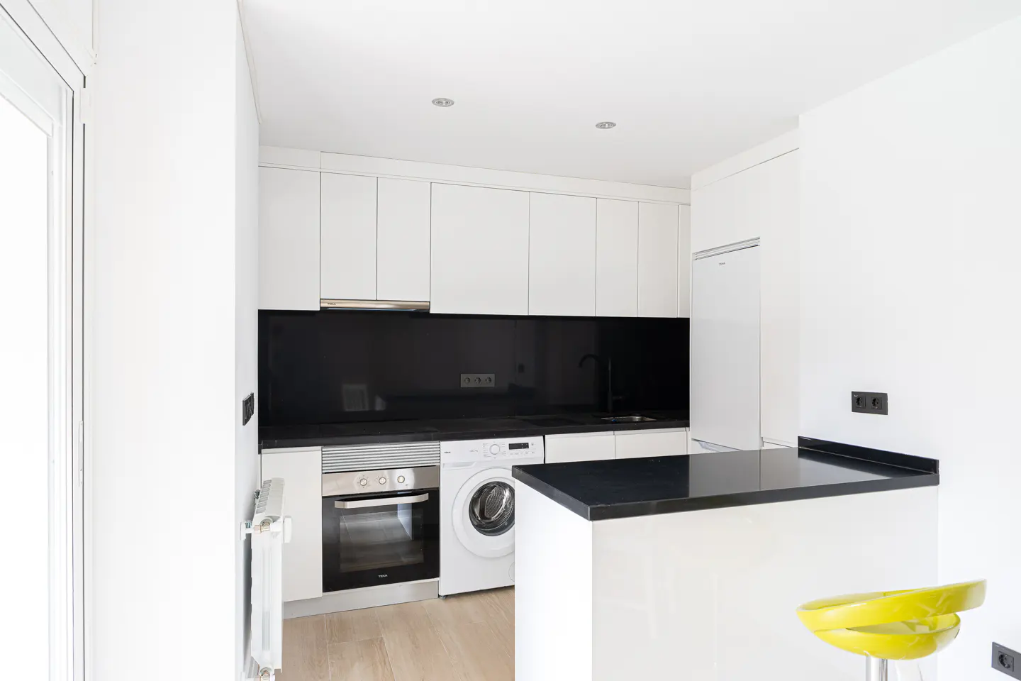 Bright, modern kitchen with white cabinets, black countertops and backsplash, oven, and washing machine. A yellow stool sits by the counter.