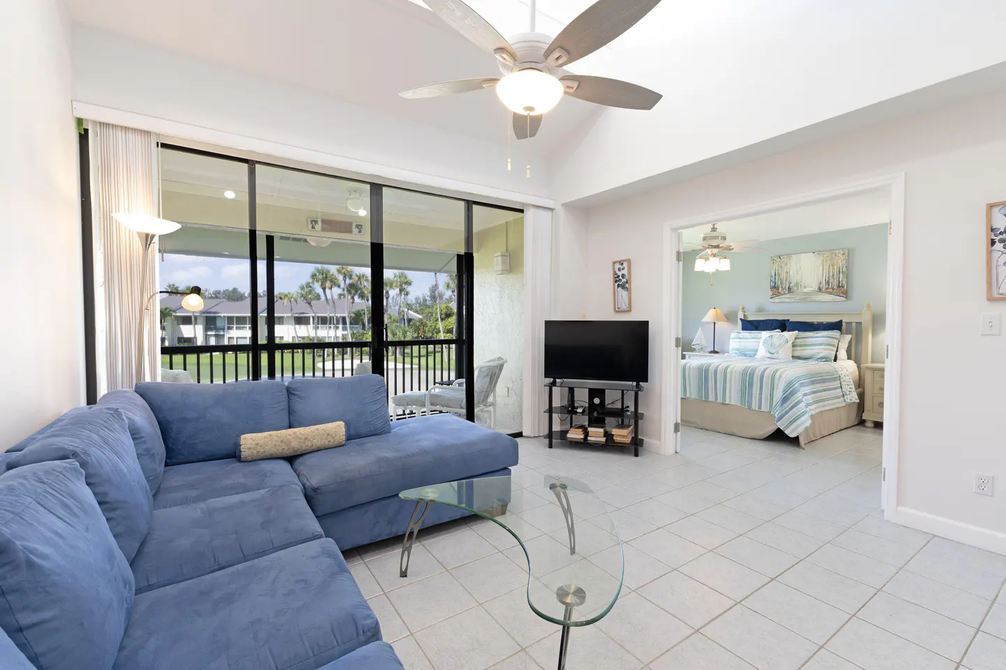 Living room with blue sectional sofa, glass table, TV, and view of bedroom with blue bedding. Balcony with palm trees visible.