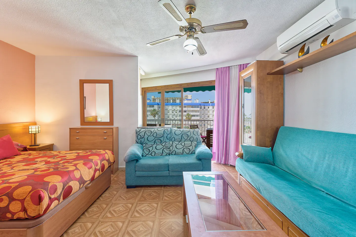 A bright studio apartment with a bed, two sofas, a ceiling fan, and a balcony with a city view.