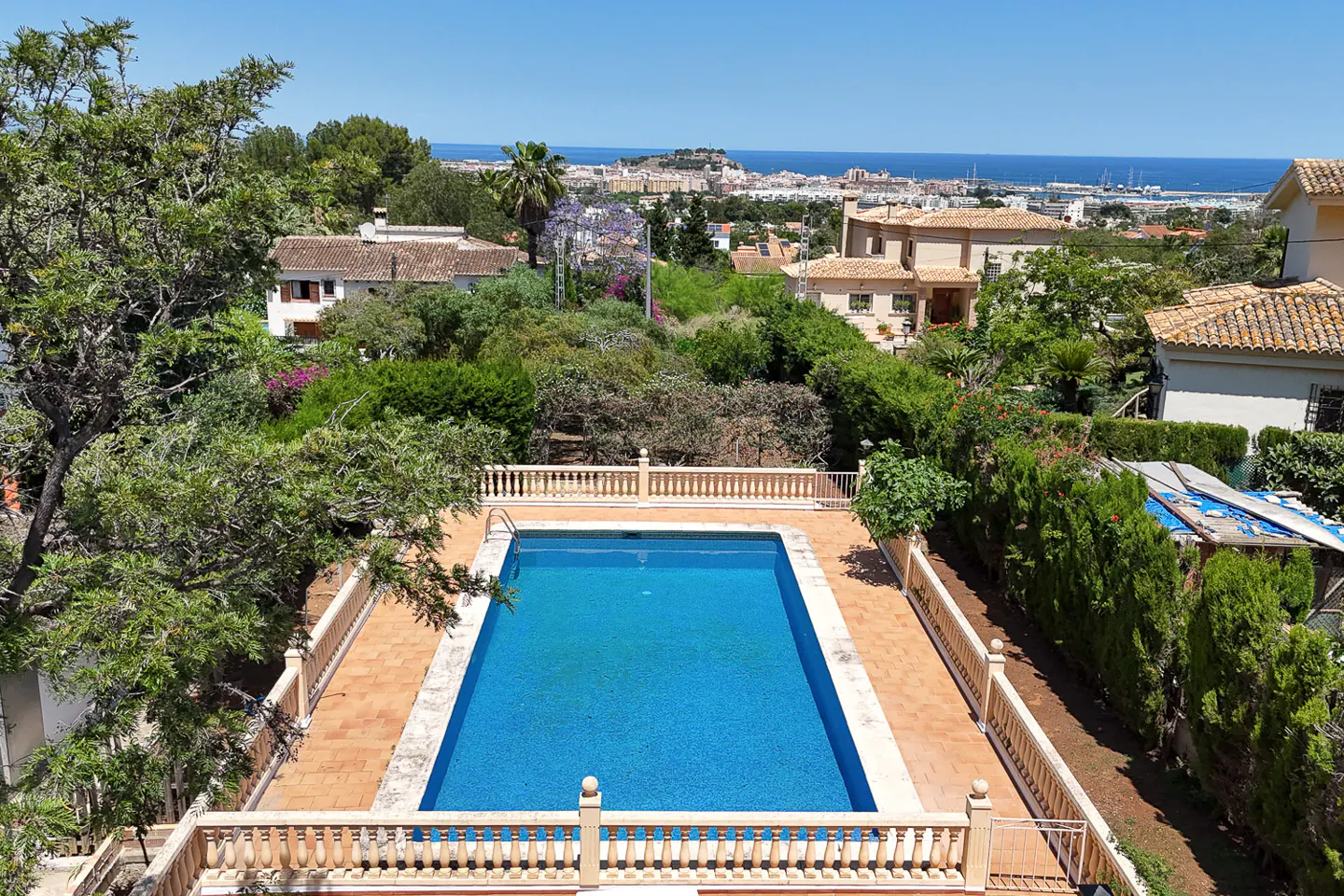 High-angle view of a rectangular blue swimming pool surrounded by a terracotta patio and balustrade, with a coastal view.