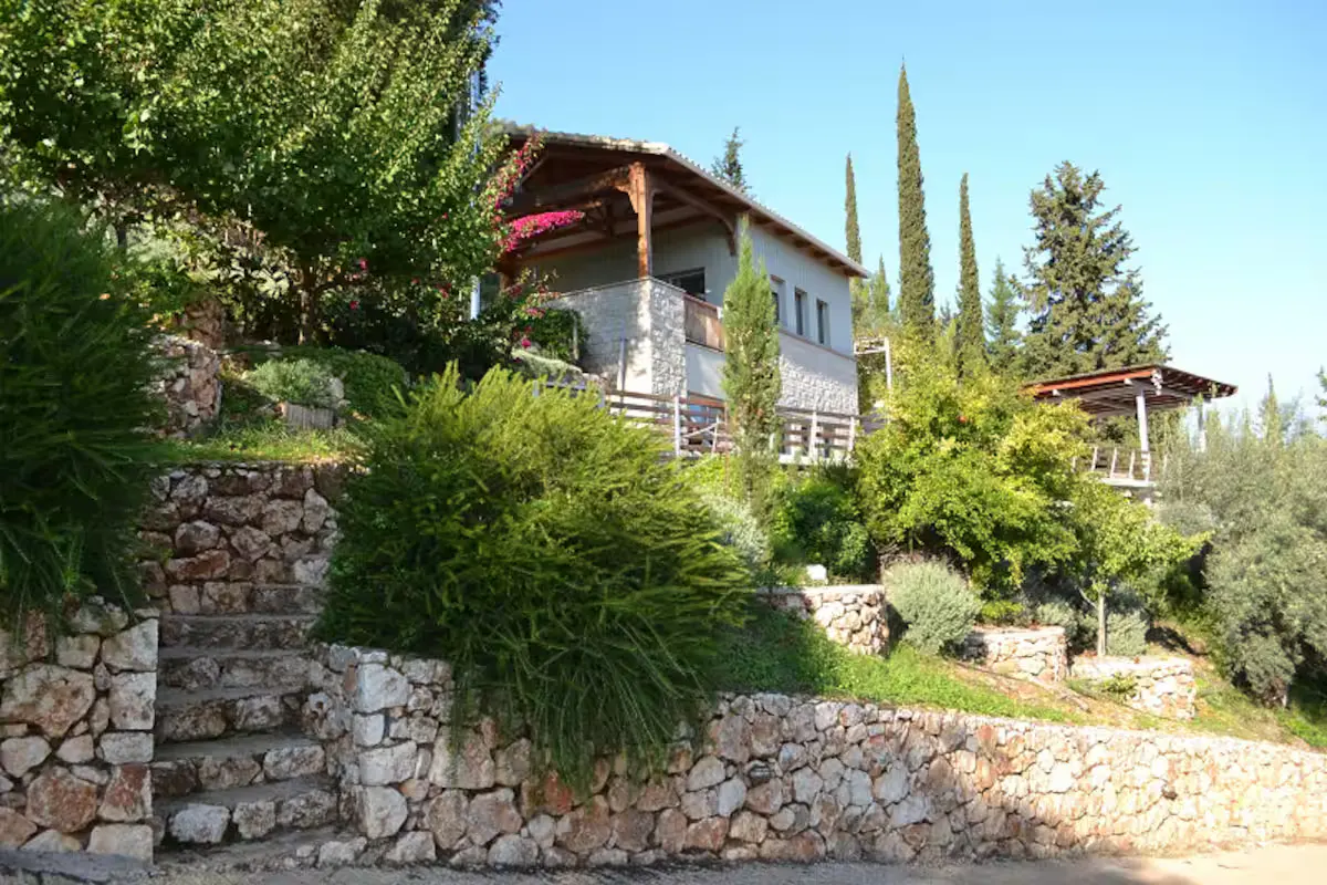 A stone house with a wooden balcony sits on a terraced hillside with stone retaining walls and lush green landscaping.