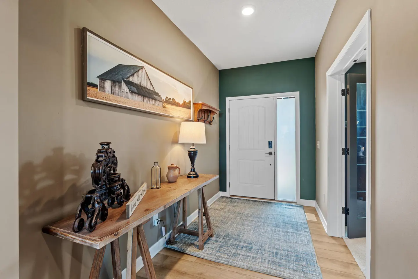 Entryway with a wooden console table, lamp, and decor. A white door and blue rug are visible. Walls are beige and green.