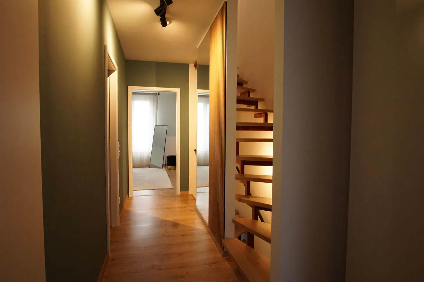 Hallway with wooden floors, green walls, and open staircase. Doors lead to rooms with natural light and a full-length mirror.