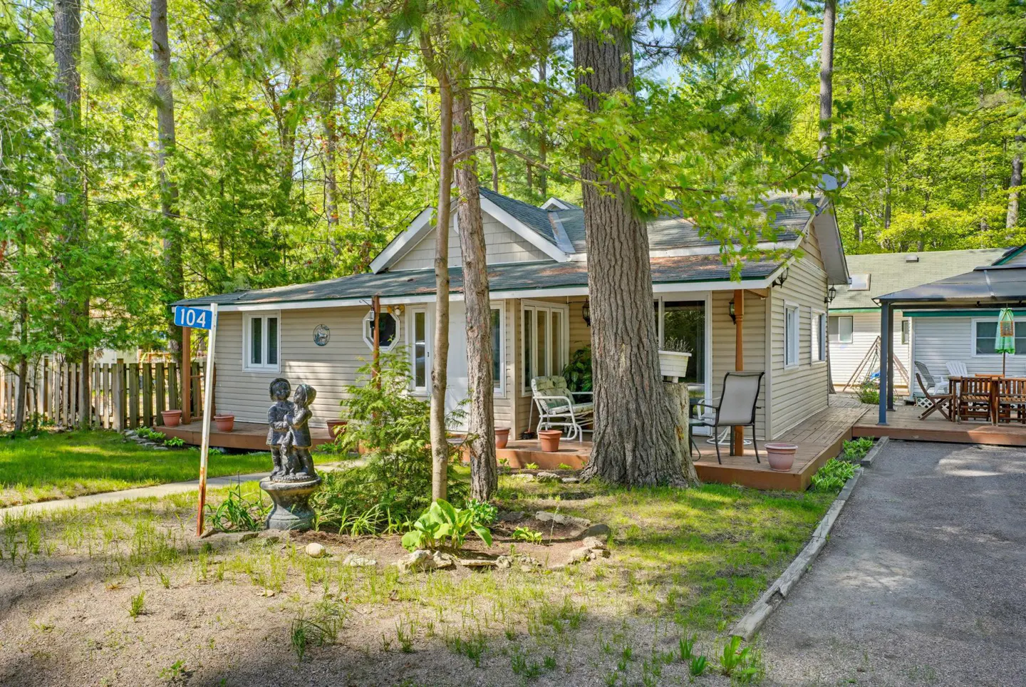 A tan cottage with a wooden deck, surrounded by lush trees, featuring a small statue of two children in the front yard.